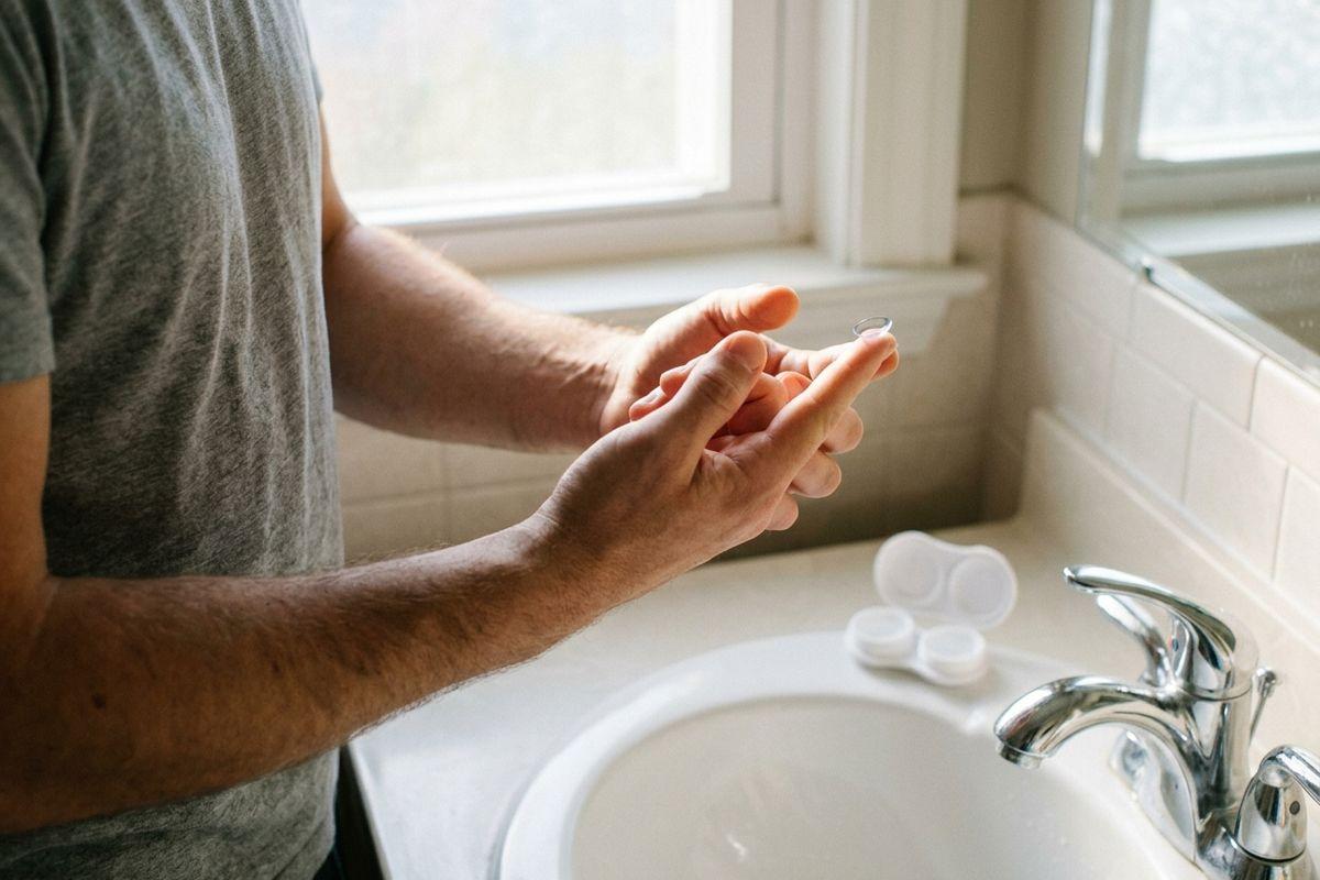 Person holding a contact lens on a fingertip at a bathroom sink, with an open contact lens case beside the faucet.